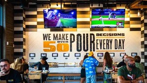 Ein Restaurant mit karierter Wand und zwei Fernsehern, die Sportveranstaltungen zeigen. Gäste speisen und stehen an der Bar.