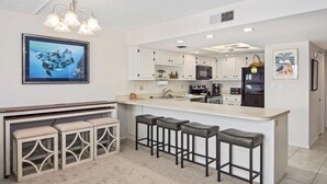 A modern kitchen with white cabinetry, a central island with bar stools, a hanging light fixture, and a framed picture on the wall.