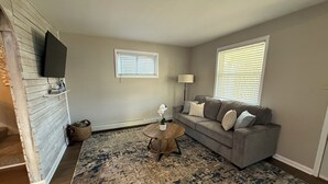 A living room with a gray sofa, a round coffee table, a patterned rug, and two windows with blinds. - The Blue Jay - Cozy 1-bedroom house in Virginia with WiFi, sleeps 4 (Virginia)