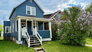 A two-story house with a front porch, a visible number '216', and a small front yard with grass and a tree with purple flowers. - The Blue Jay - Cozy 1-bedroom house in Virginia with WiFi, sleeps 4 (Virginia)