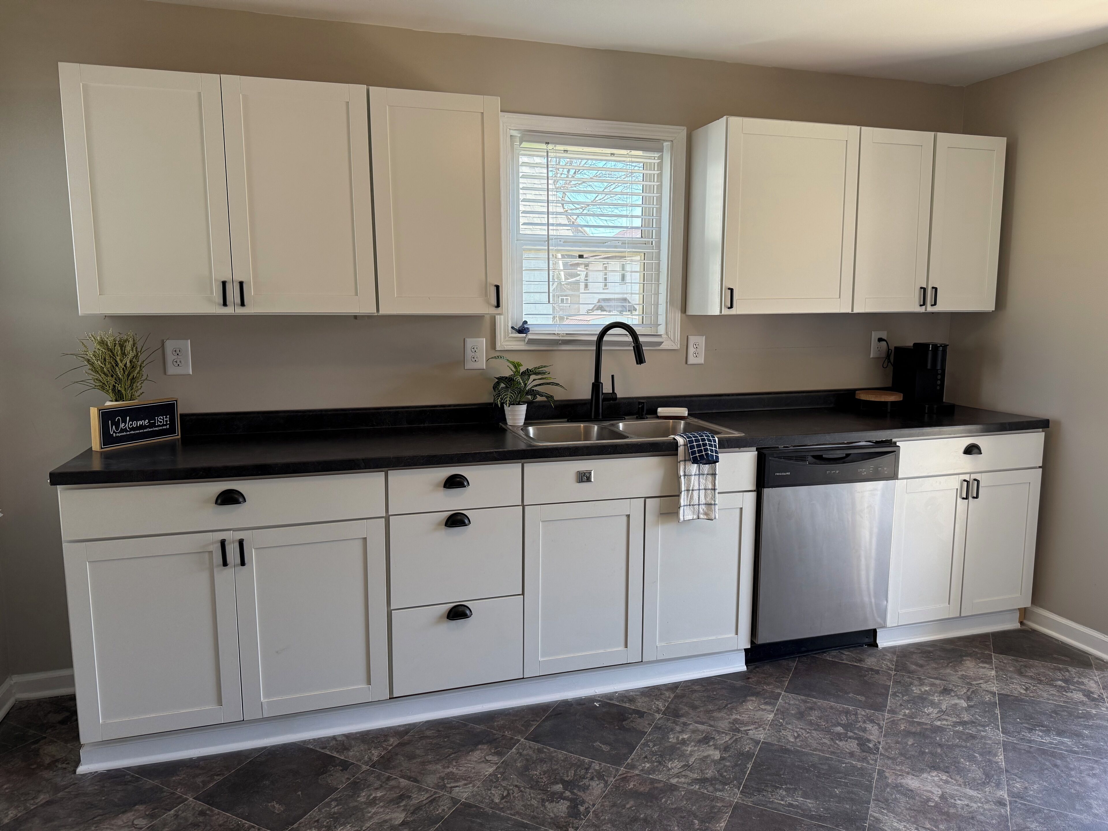 A kitchen with white cabinets, a black countertop, a sink, and a window with blinds.