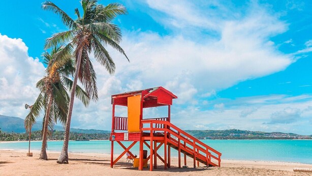 Ein Strand mit einem roten Rettungsturm, Palmen und klarem, blauem Wasser.