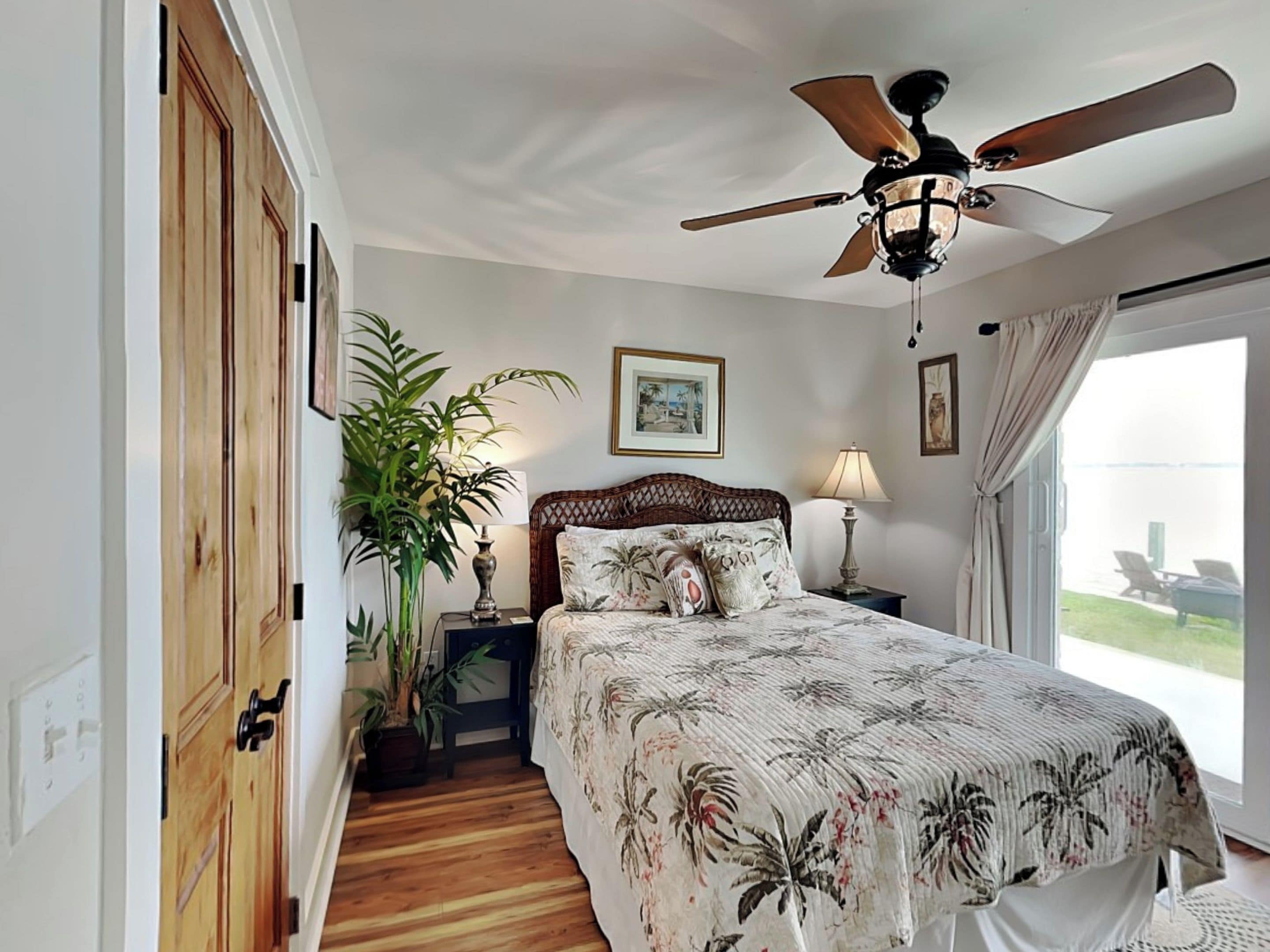 A bedroom with a wooden floor, a bed with a floral bedspread, a ceiling fan, and a view of the outdoors through a window.