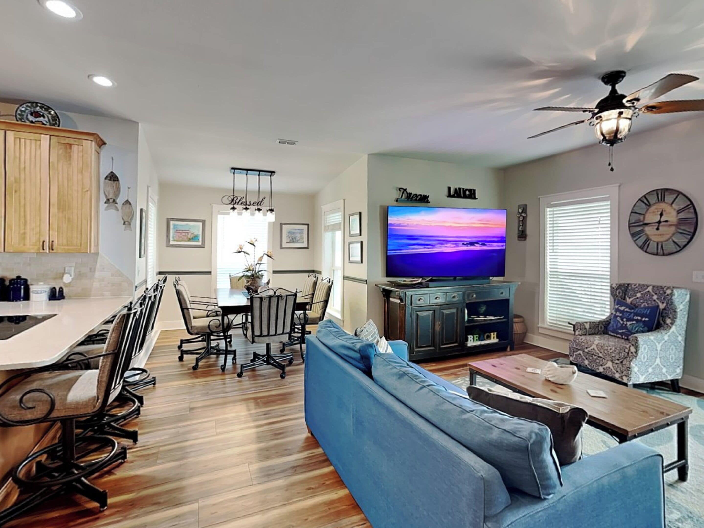 A modern living room with a blue sofa, a coffee table, and a television. There is a dining area with a table and chairs, and a kitchen with wooden cabinets.
