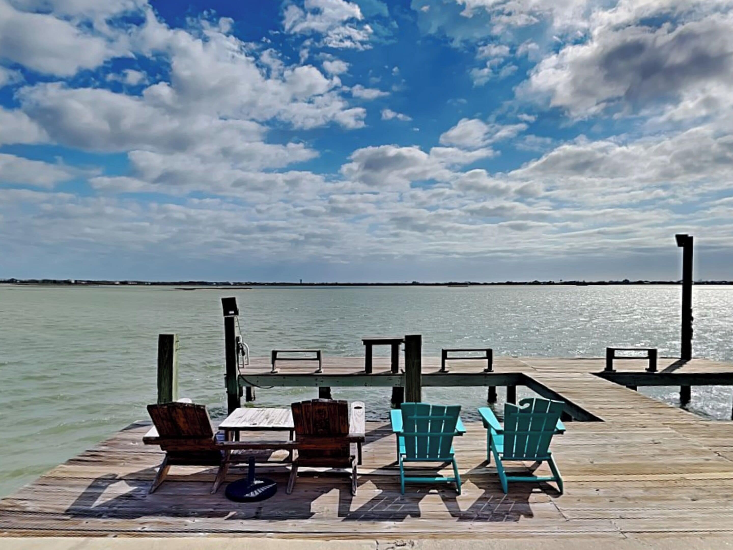A wooden pier with several Adirondack chairs and a table, overlooking a calm body of water under a partly cloudy sky.