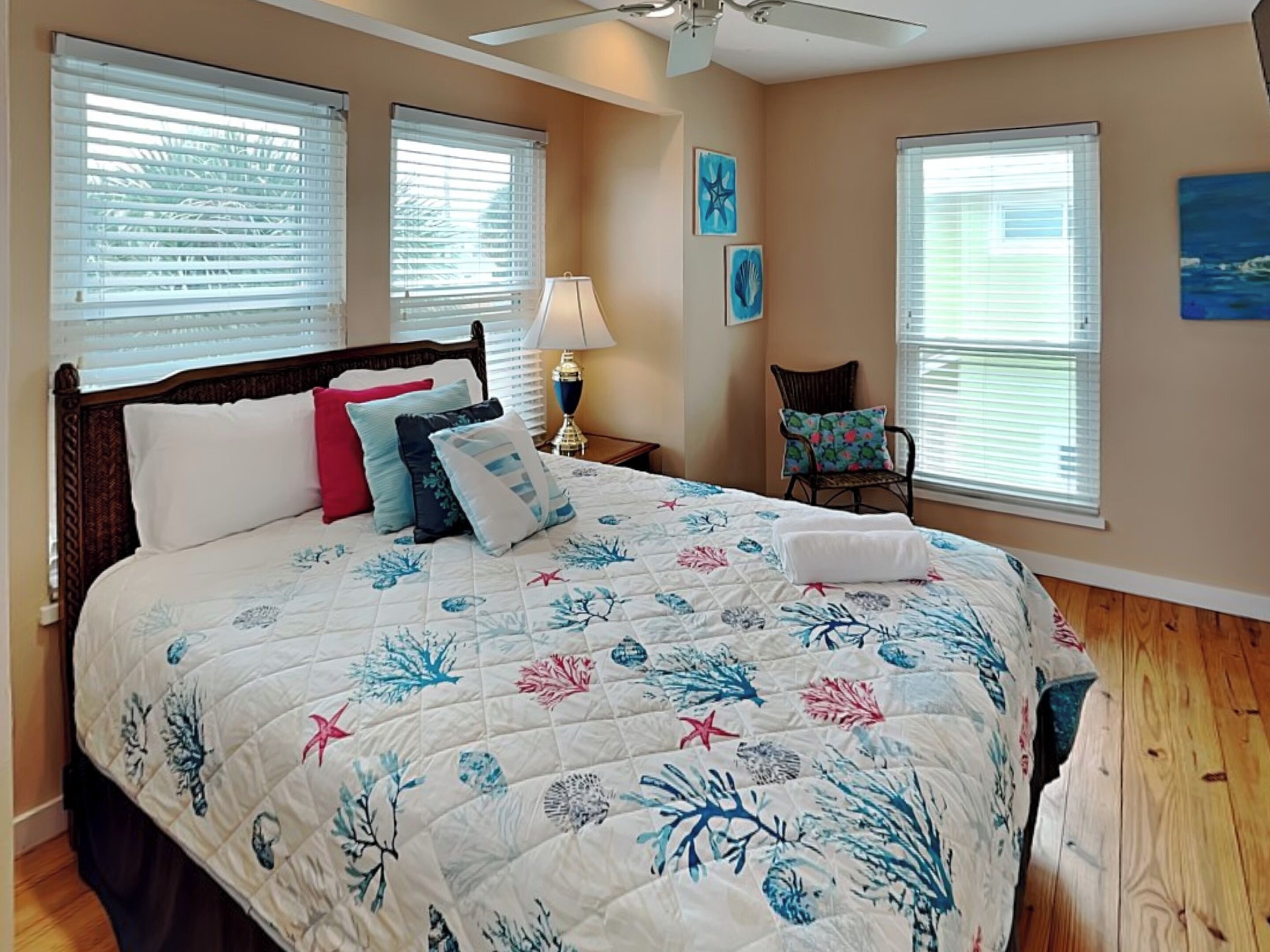 A bedroom with a bed, two windows, a ceiling fan, a chair, and artwork on the walls.