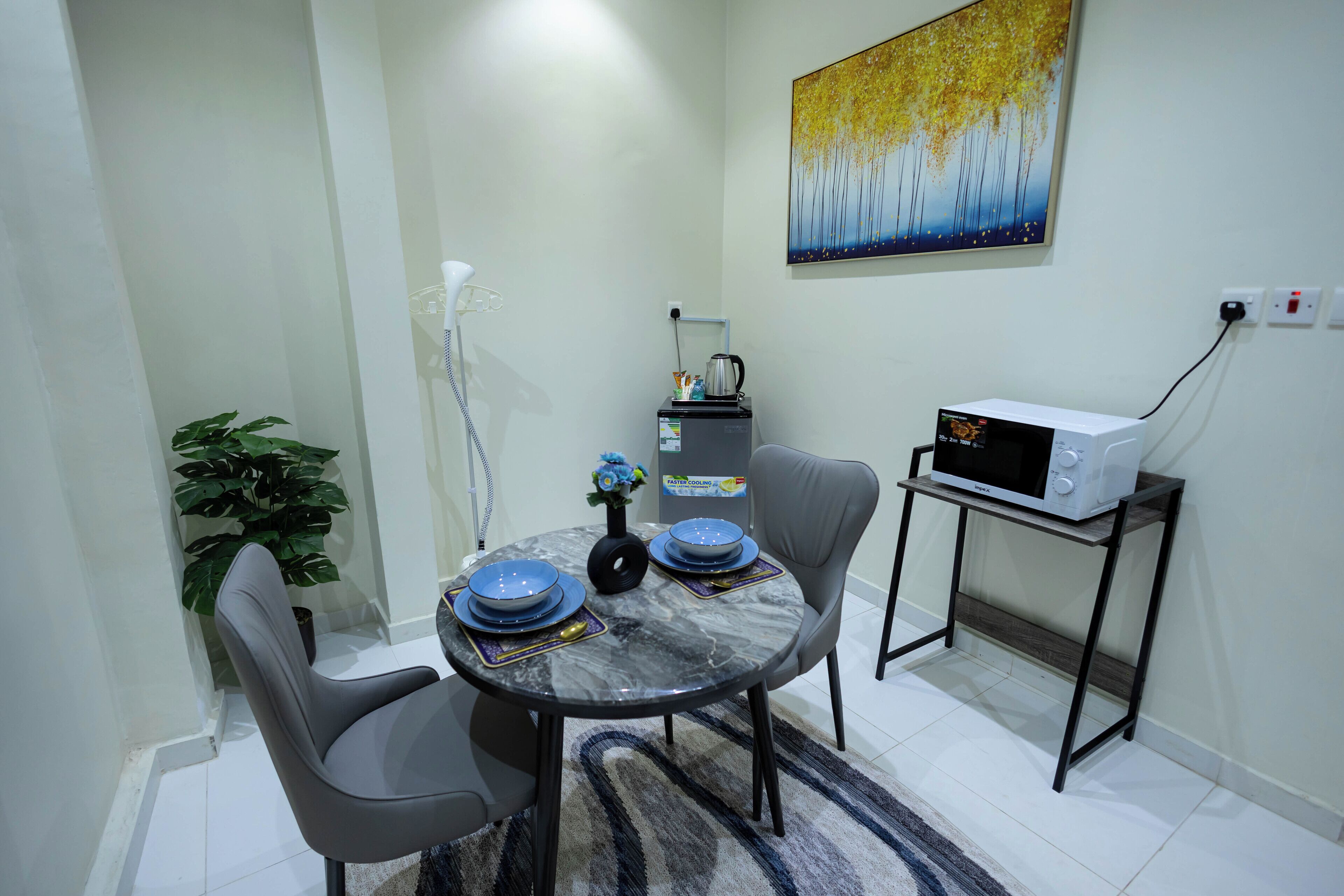 A dining area with a round table, two chairs, a microwave on a small table, a plant, and a painting on the wall.