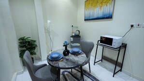 A dining area with a round table, two chairs, a microwave on a small table, a plant, and a painting on the wall. - Centrally Located Self Checkin Luxury  Apartment (Riyadh)