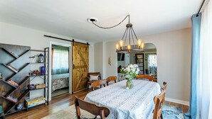 A dining area with a wooden table, chairs, and a chandelier. There is a bookshelf with books and decorative items, and a wooden door leading to another room.