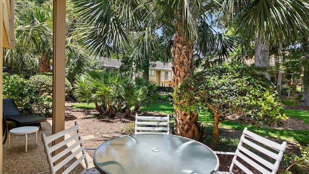 Une terrasse dotée d’une table ronde en verre et de chaises en métal blanc, entourée d’une végétation luxuriante et de palmiers.