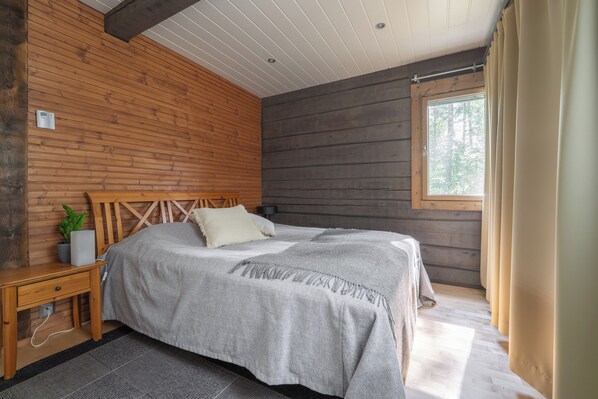 Un dormitorio con cabecera de madera, cama con manta gris, mesita de noche con lámpara y ventana con cortinas.