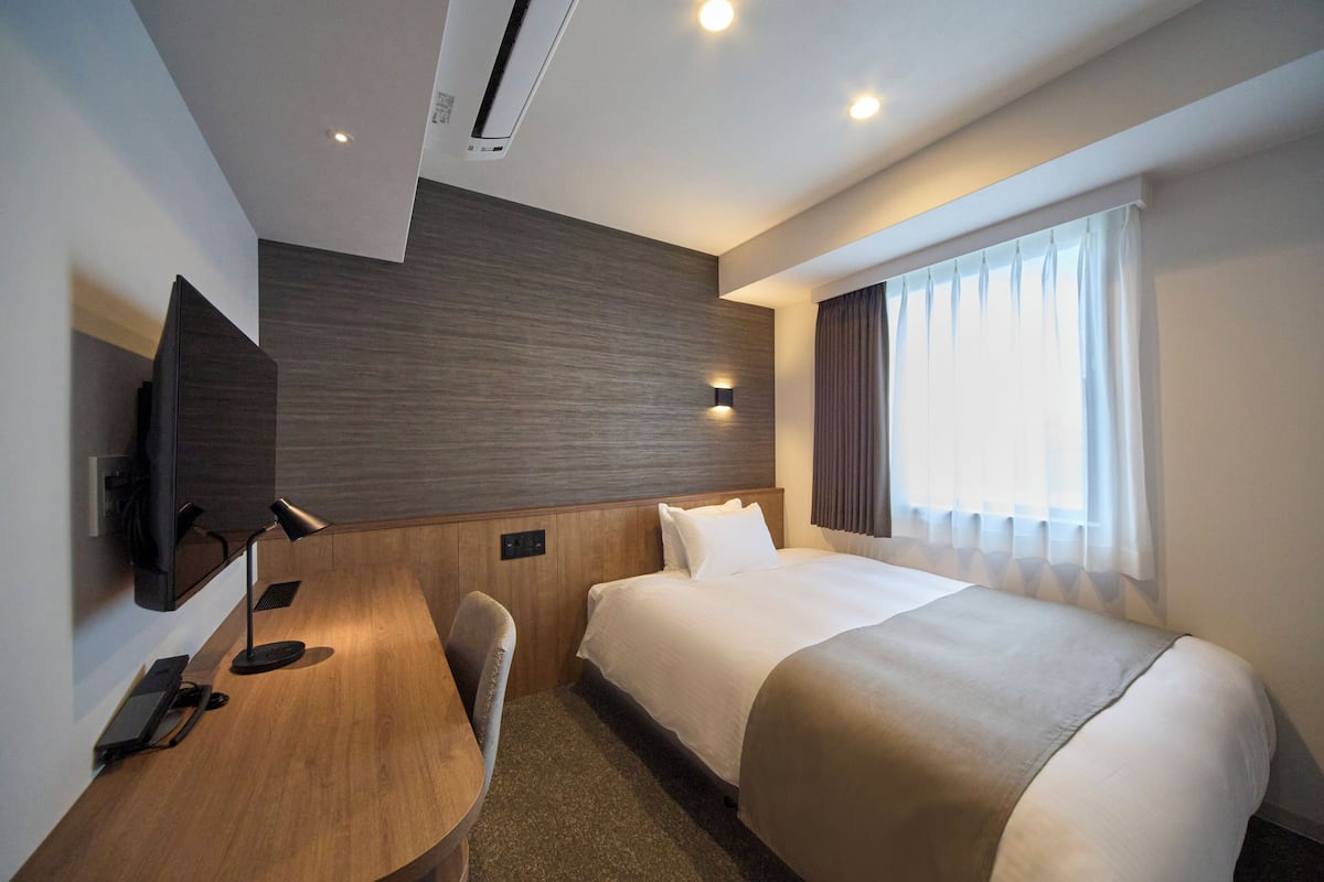 A hotel room with a bed, a desk with a chair, a flat-screen TV, and a window with curtains.