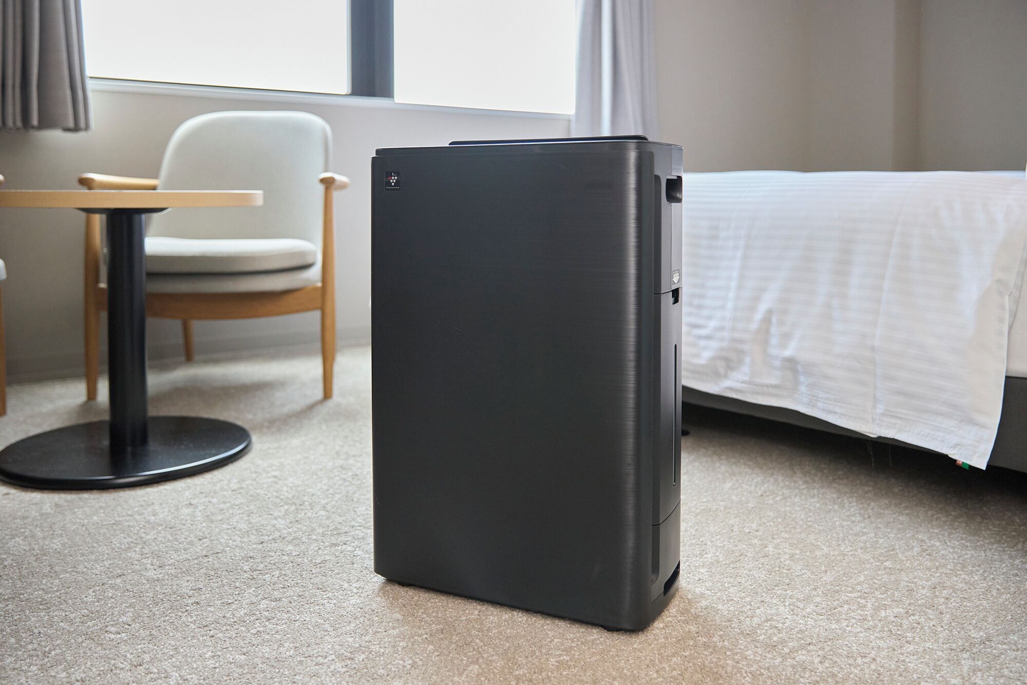 A compact, dark-colored refrigerator in a hotel room with a bed, a chair, and a small table.