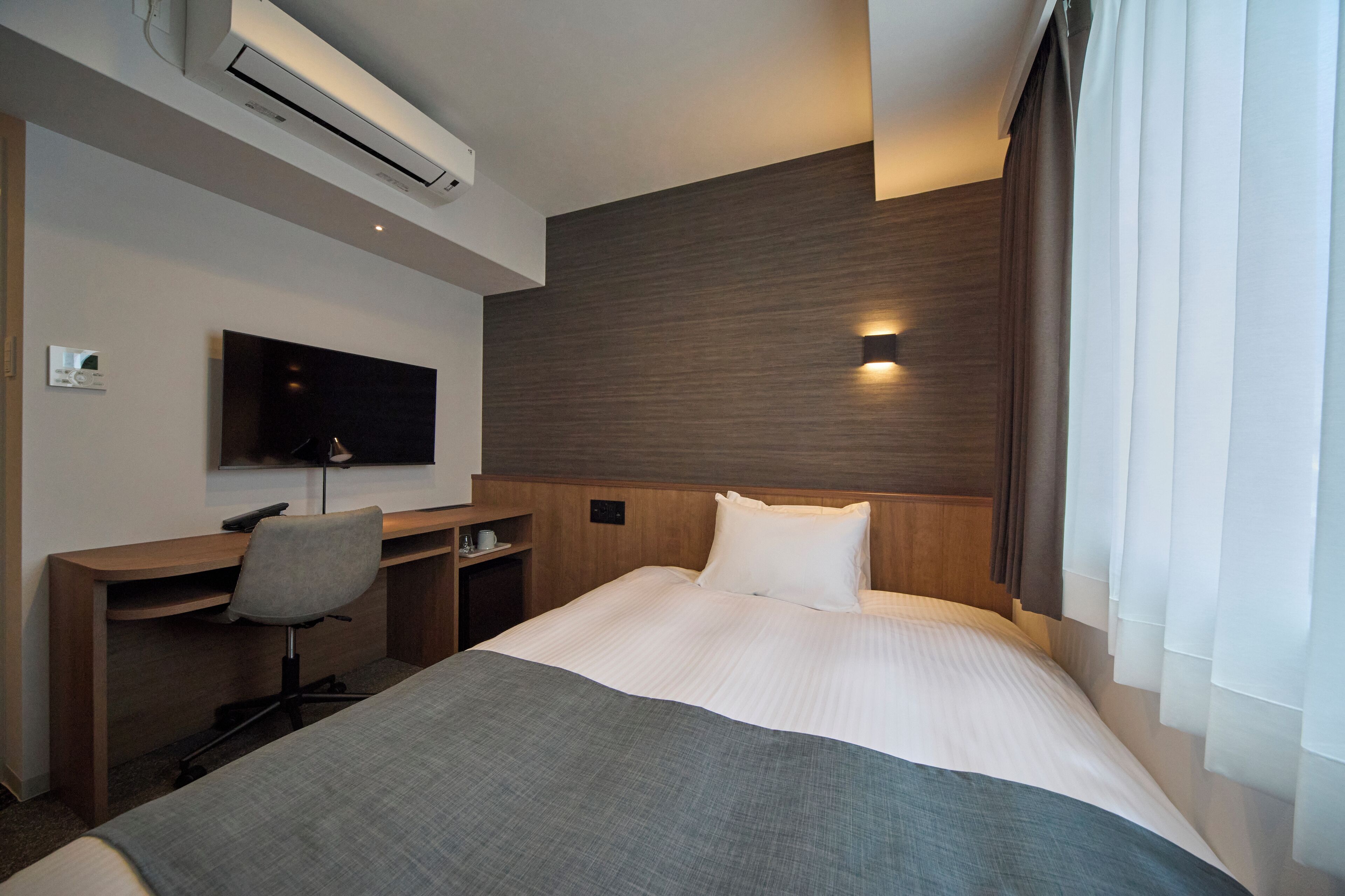A hotel room with a bed, a desk, a chair, a TV, and an air conditioning unit.