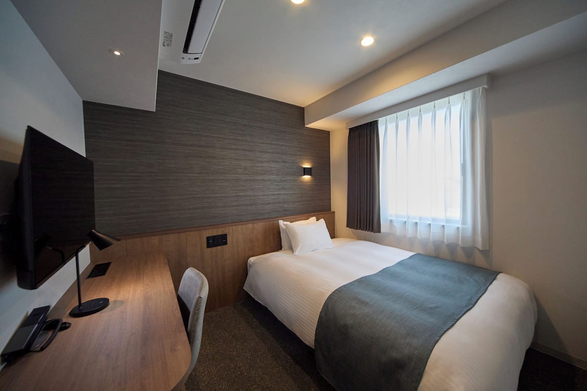 A hotel room with a bed, a desk with a telephone, a television, and a window with curtains.