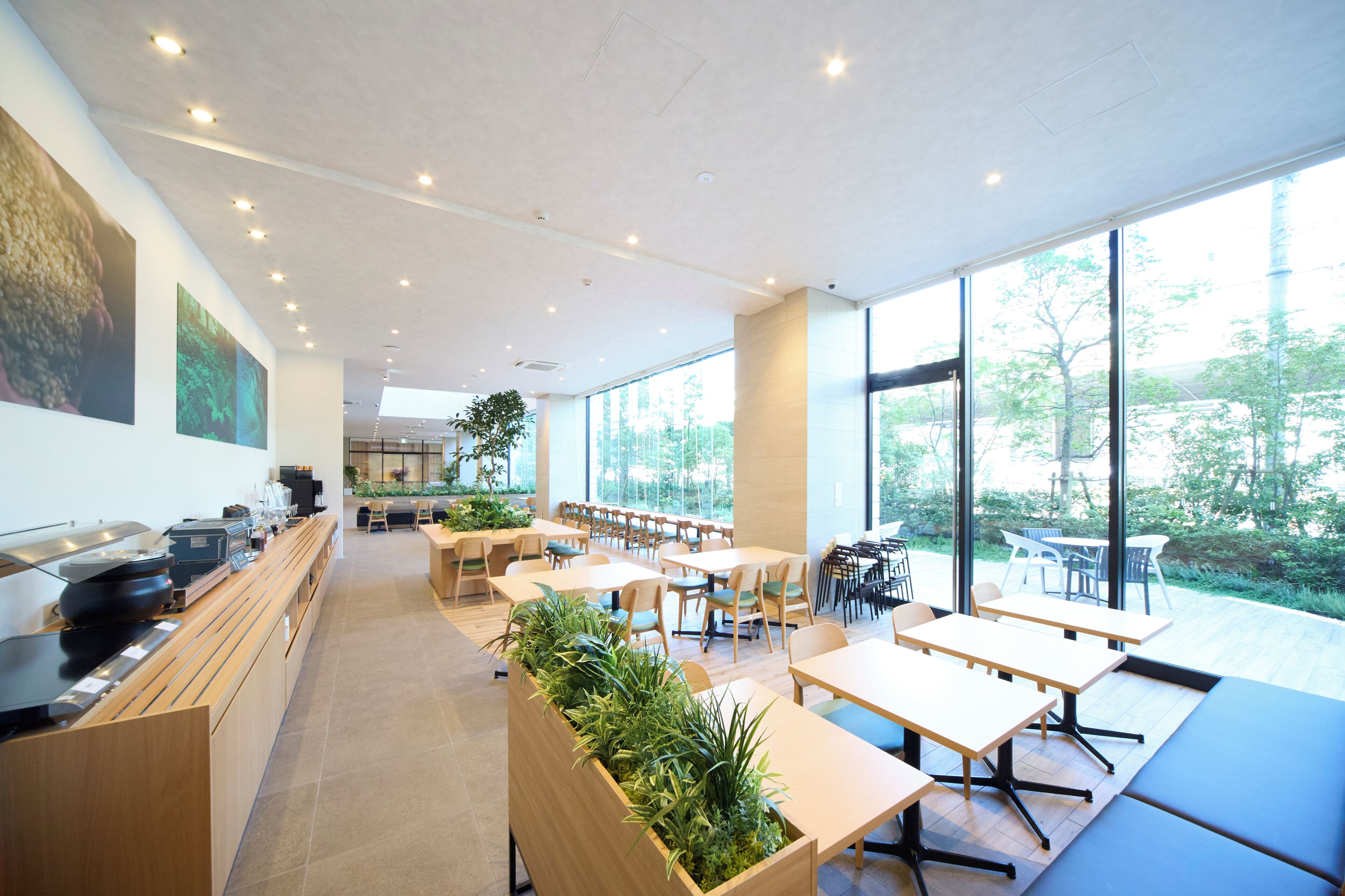A modern cafe with wooden tables and chairs, large windows offering a view of greenery, and a counter with coffee machines.