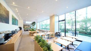 A modern cafe with wooden tables and chairs, large windows offering a view of greenery, and a counter with coffee machines.
