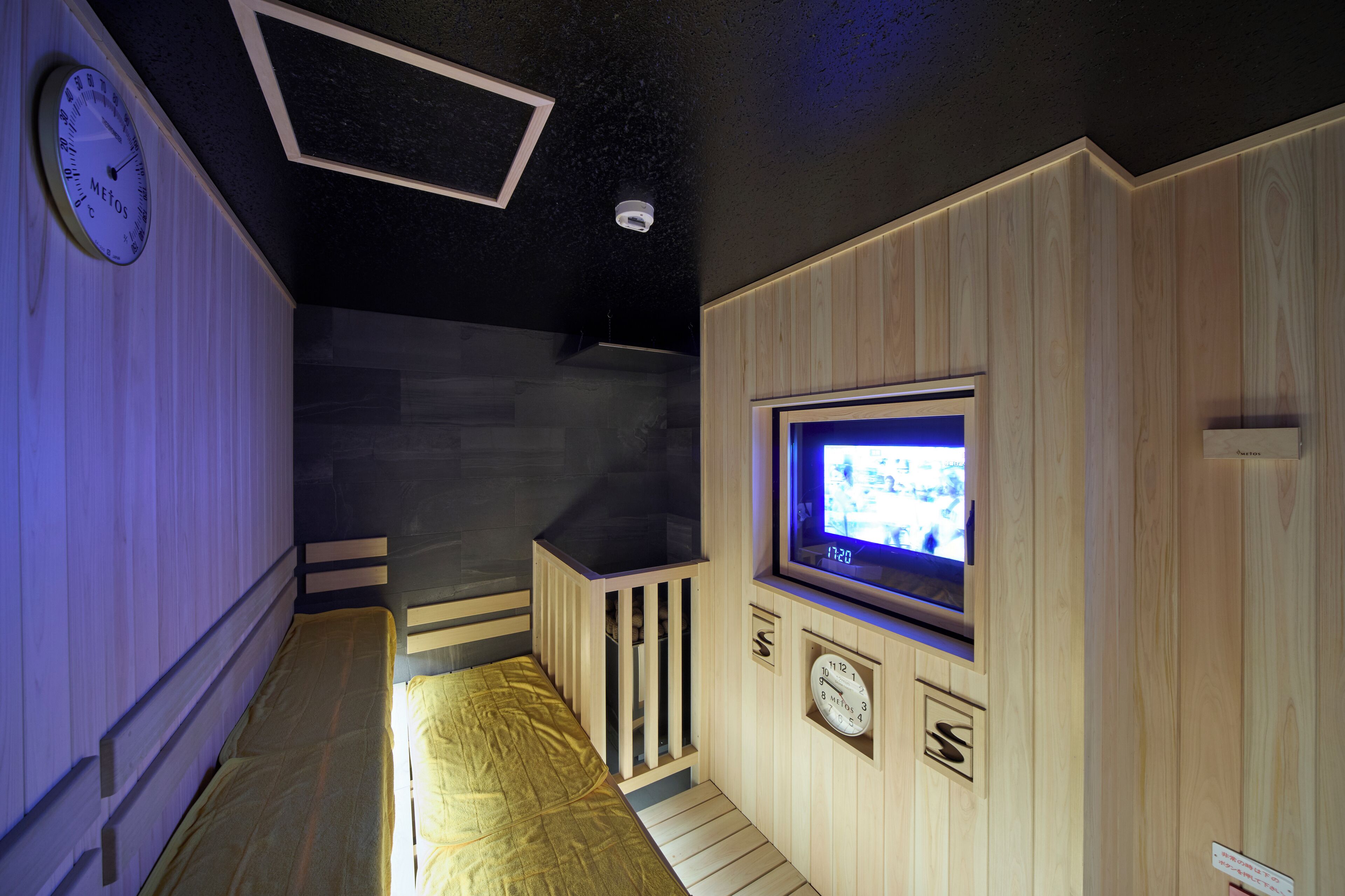 A room with a bed, a television mounted on the wall, a clock, and a small window.