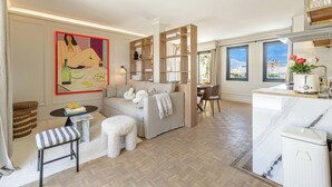 A modern living room with a sofa, ottoman, and a coffee table. There is a dining area with a table and chairs. The room has large windows and a view of the city. - Exceptional Penthouse, 2 BDR Suites, 3 spacious terraces, Sea view, Elevator, AC (Nice)