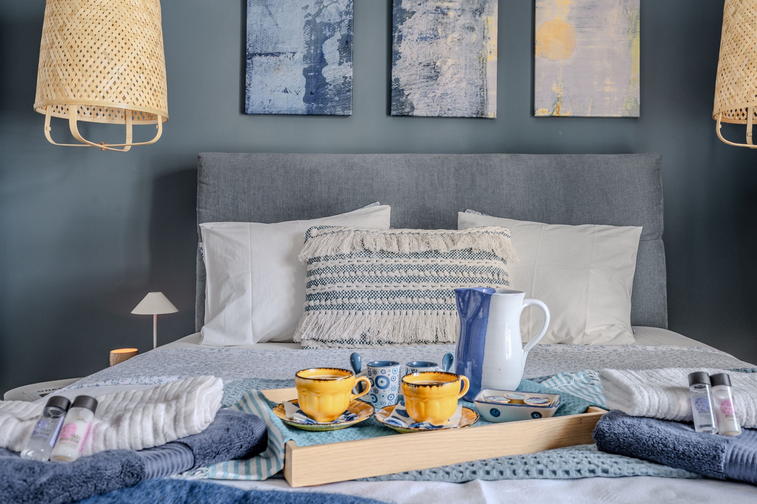 Una cama bien tendida con un cabecero gris, dos almohadones decorativos, una bandeja con tazas y una jarrera, y una lámpara de mesa pequeña.