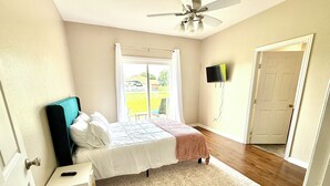 A bedroom with a bed, a nightstand, a ceiling fan, a window with curtains, a TV mounted on the wall, and a door.