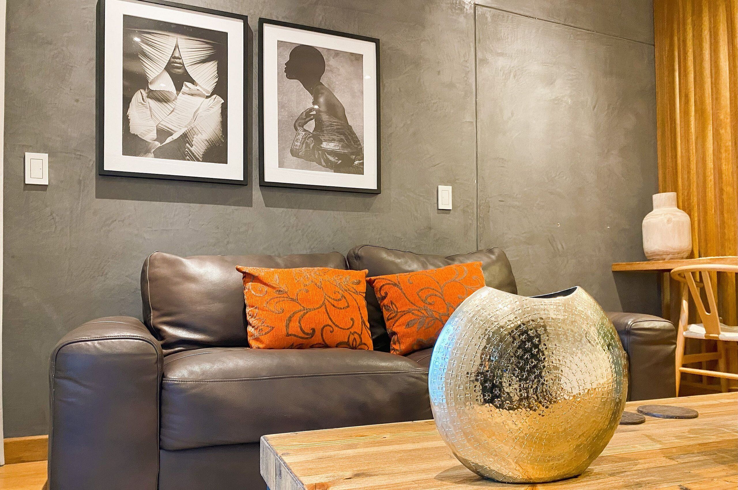 A living room with a brown leather sofa, orange patterned cushions, and a decorative vase on a wooden table. Two framed artworks are hung on the wall.