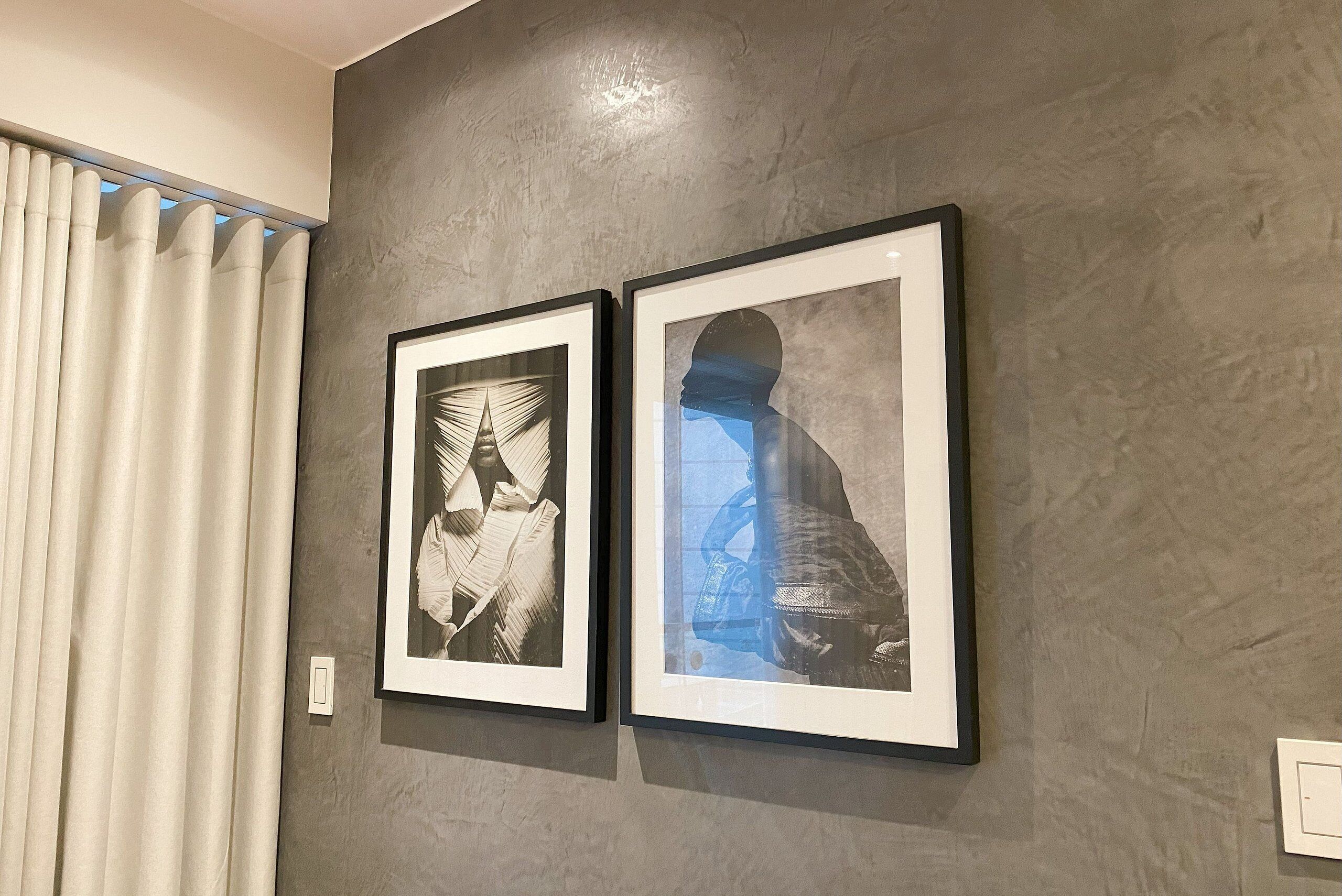 Zwei gerahmte Kunstwerke an einer strukturierten Wand, ein Fenster mit weißen Vorhängen und ein Lichtschalter.