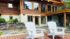Un terrazzo in legno con due poltrone Adirondack bianche, un muro in pietra e una casa a due piani con ampie finestre e un balcone.