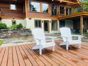 Eine Holzterrasse mit zwei weißen Adirondack-Stühlen, einer Steinmauer und einem zweigeschossigen Haus mit großen Fenstern und einem Balkon.