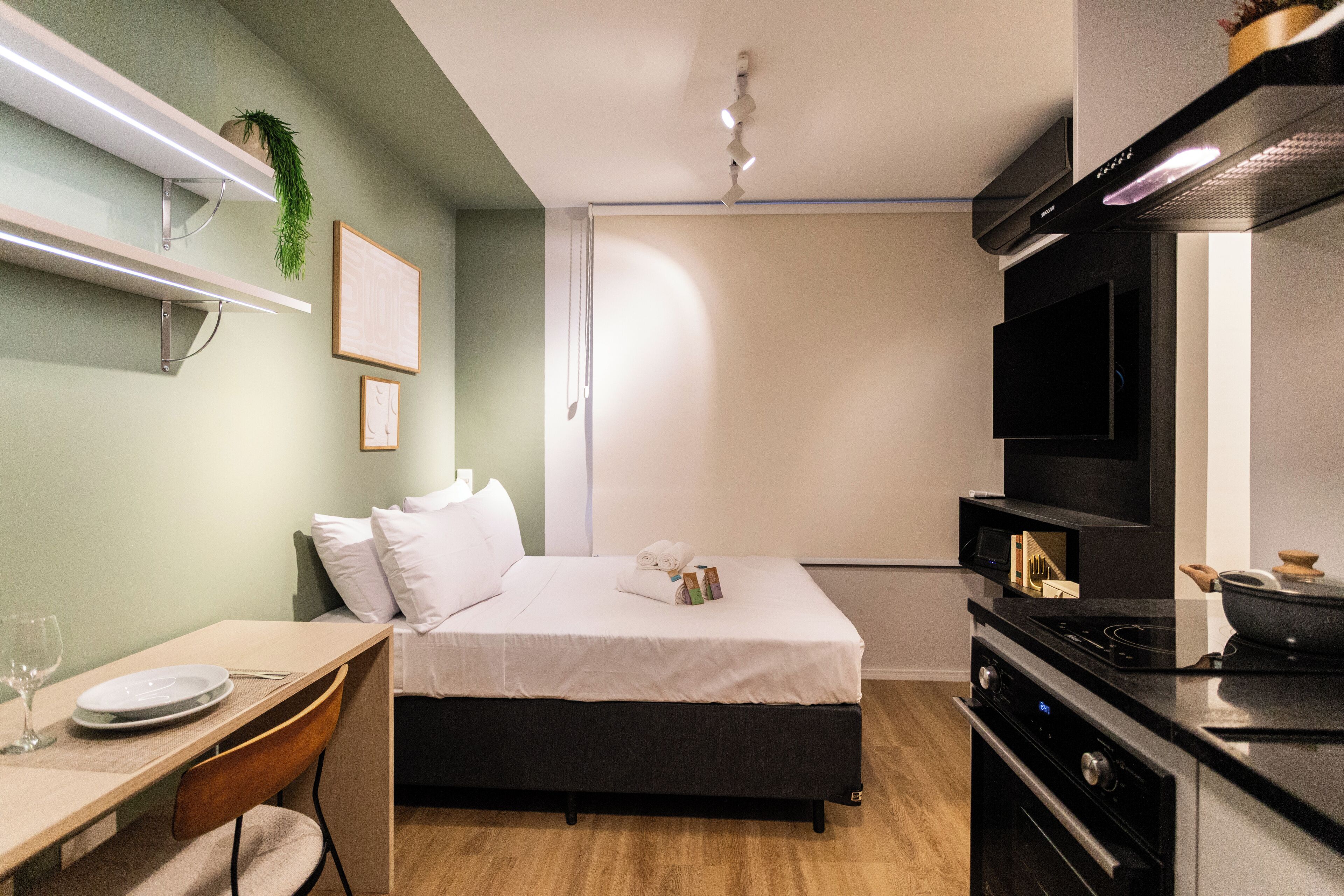 A modern hotel room with a bed, a desk with a chair, a television, and a kitchen area with a stove.