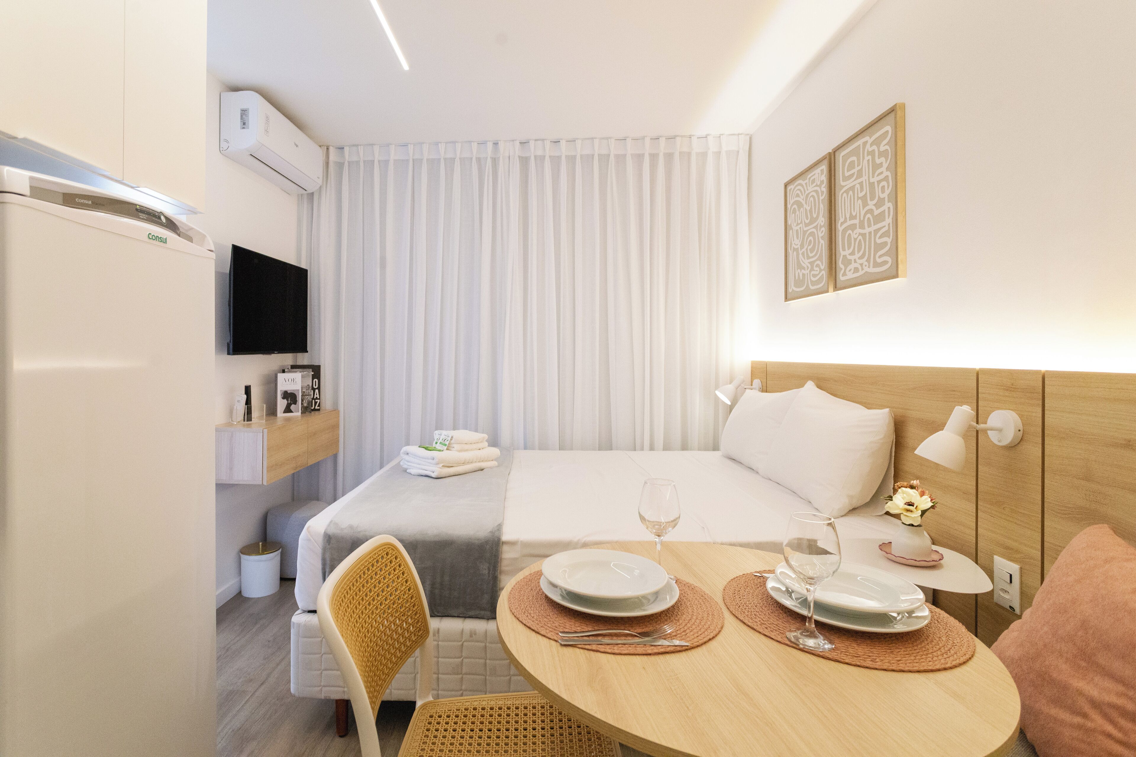 A modern hotel room with a bed, bedside table with a lamp, a TV mounted on the wall, a dining area with a table and chairs, and a window with curtains.