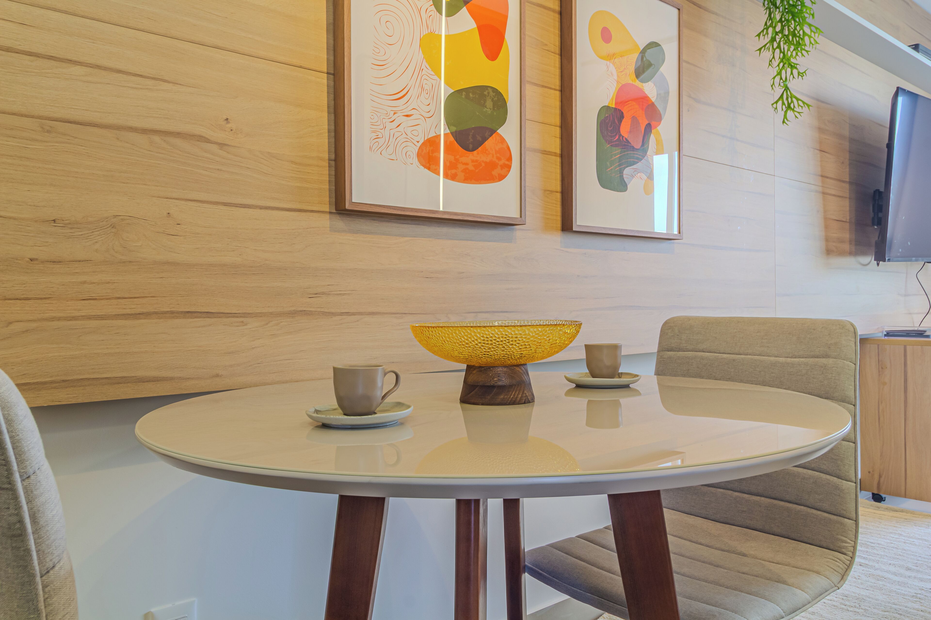 A round table with a white top, two chairs, a yellow woven bowl, and two cups on a wooden floor with a wooden wall and two framed artworks.