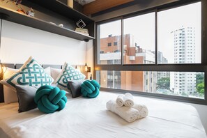 Hotellihuone, jossa on suuri ikkuna, valkoisilla lakanoiden peitetty sänky, turkoosinsiniset ja harmaat tyynyt sekä näkymä kaupungin rakennuksiin.