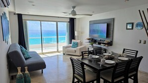 Un salon moderne avec un balcon donnant sur l’océan, un coin repas avec une table dressée pour deux personnes et un téléviseur à écran plat fixé au mur.