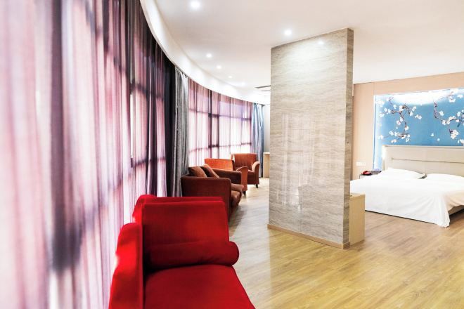 A hotel room with a curved wall, a bed, a seating area with red chairs, and a large window with a floral design.