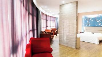 A hotel room with a curved wall, a bed, a seating area with red chairs, and a large window with a floral design.