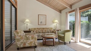 Een woonkamer met een bank, fauteuil en salontafel, met een groot raam en een plafond met zichtbare balken.