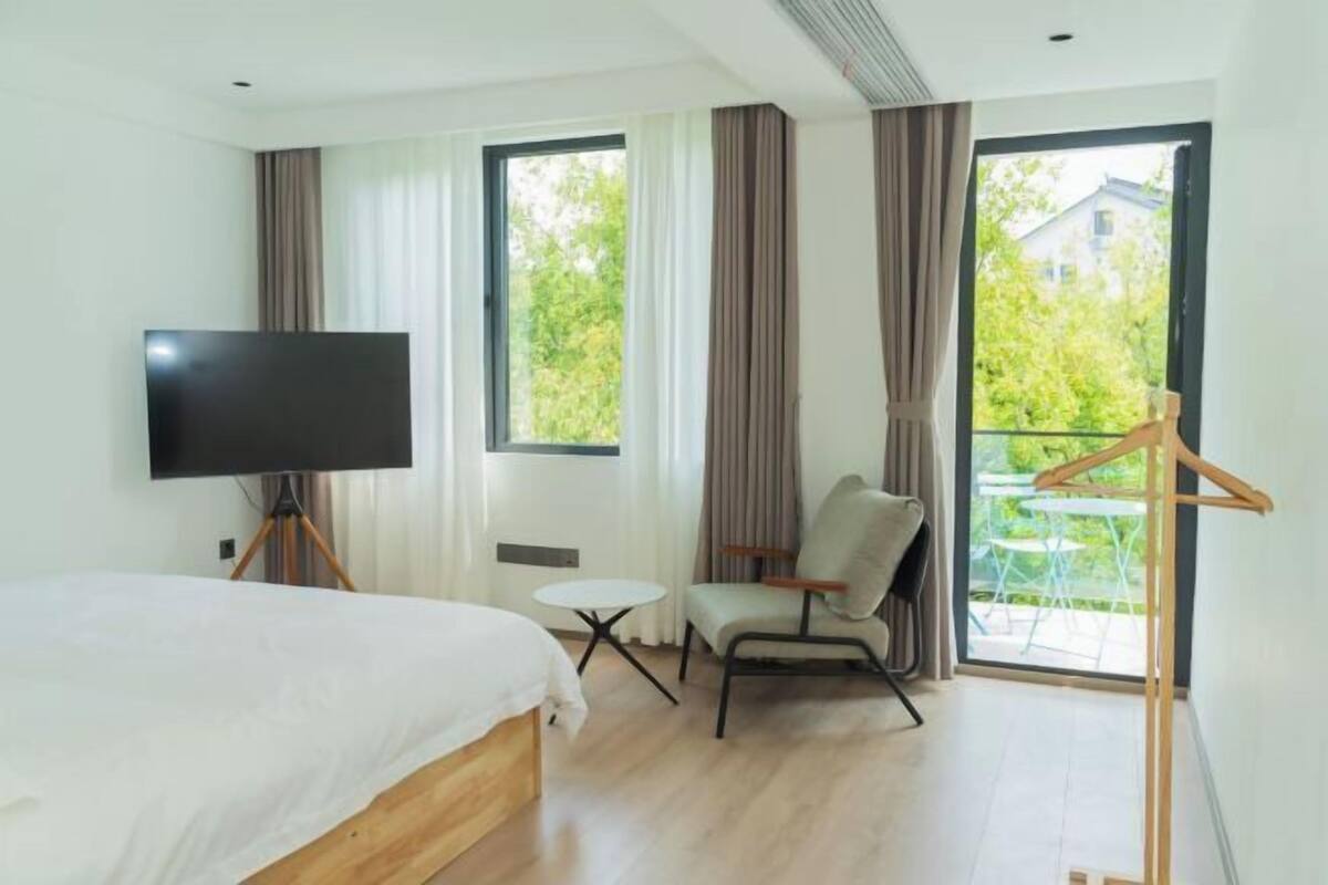 Une chambre d’hôtel moderne équipée d’un lit, d’une télévision, d’une chaise, d’une petite table et d’un balcon donnant sur un paysage verdoyant.