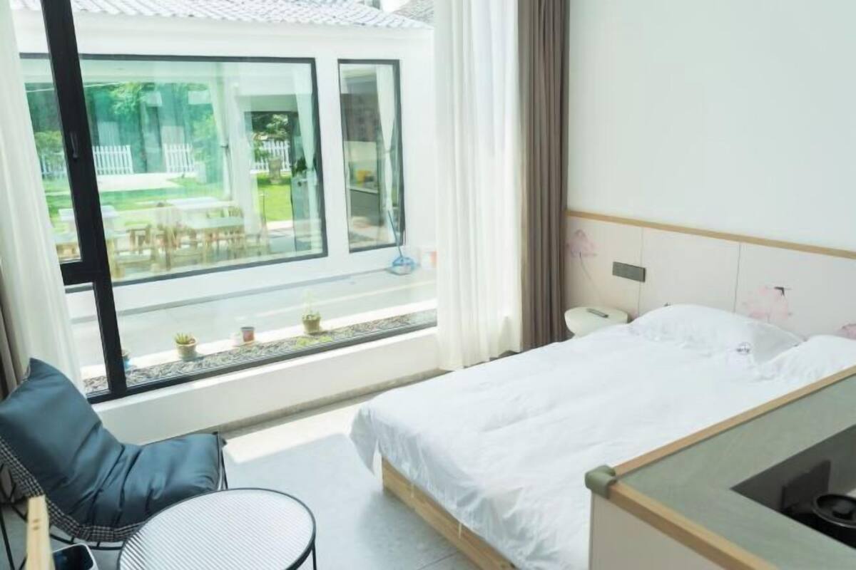Une chambre d’hôtel avec un lit, une chaise, une petite table et une vue sur un jardin, visible à travers une grande fenêtre.