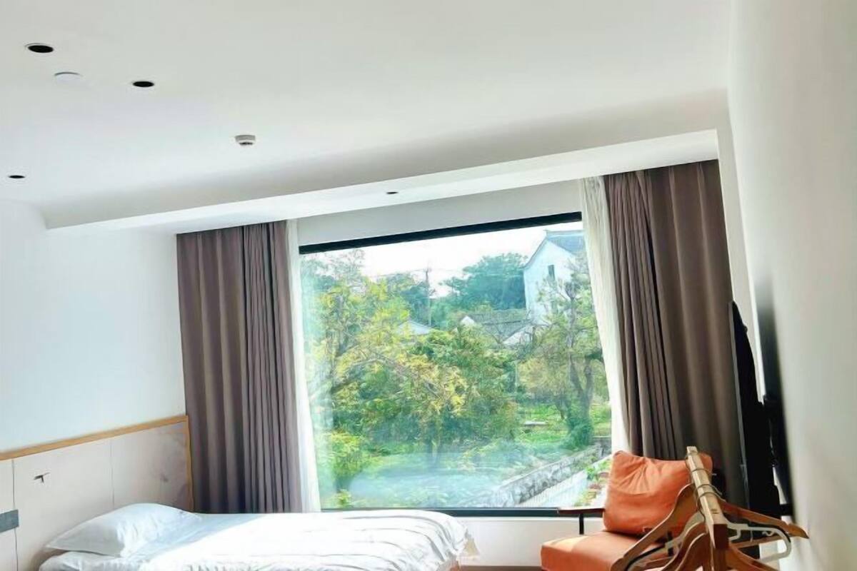 Une chambre d’hôtel moderne dotée d’une grande fenêtre, d’un lit et d’un coin salon avec un fauteuil et un cintre en bois.