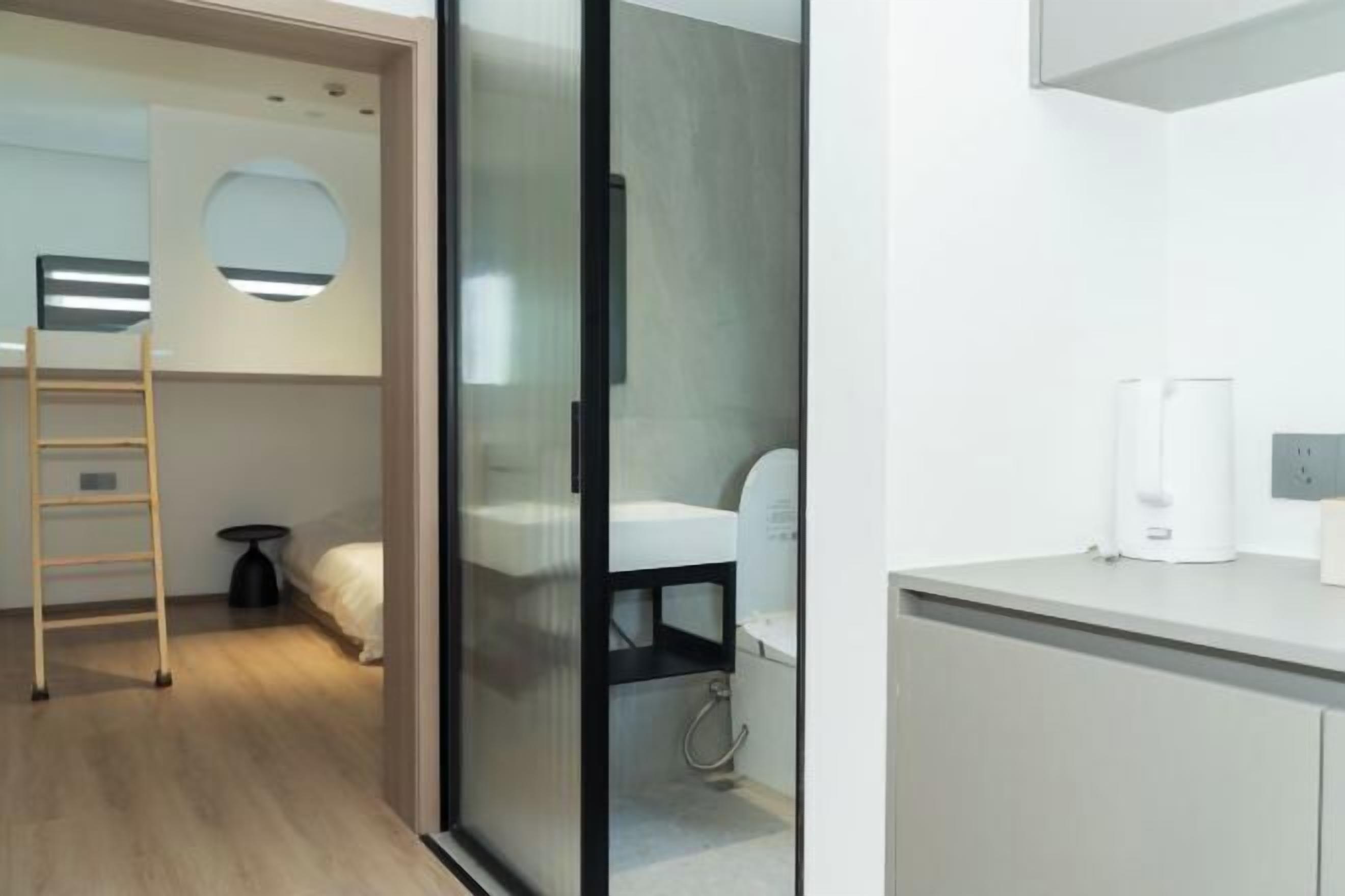 A modern hotel room with a glass partition, a wooden ladder, a white bed, a small round mirror, and a white sink.