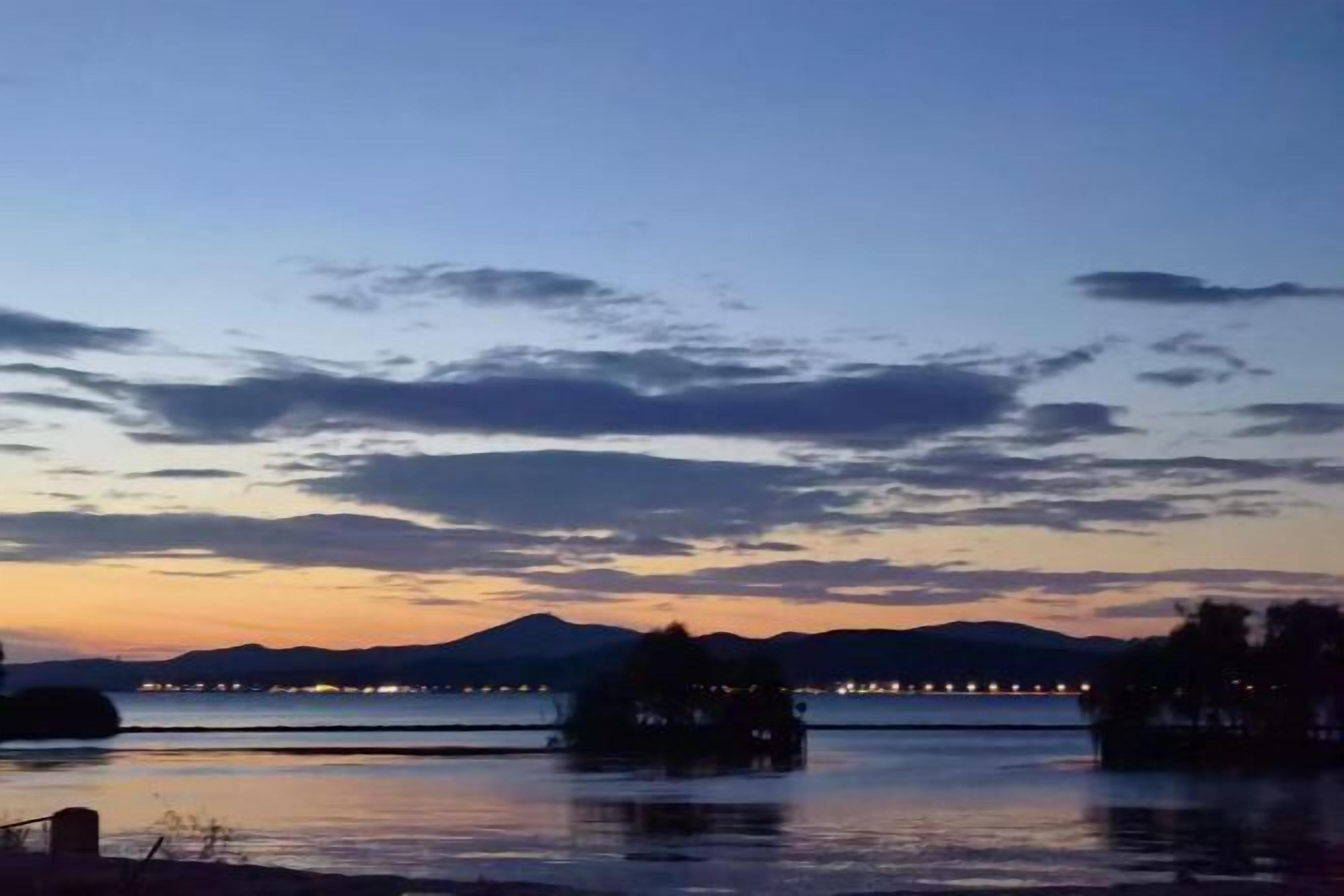 Une vue paisible sur un lac au crépuscule, avec une chaîne de montagnes lointaine, une structure flottante et un pont illuminé.