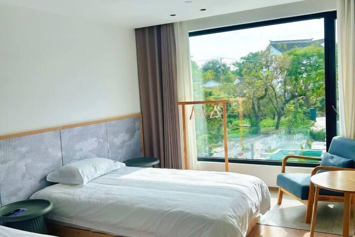 Une chambre d’hôtel avec un lit, une chaise, une petite table et une grande fenêtre donnant sur un paysage verdoyant et des bâtiments.