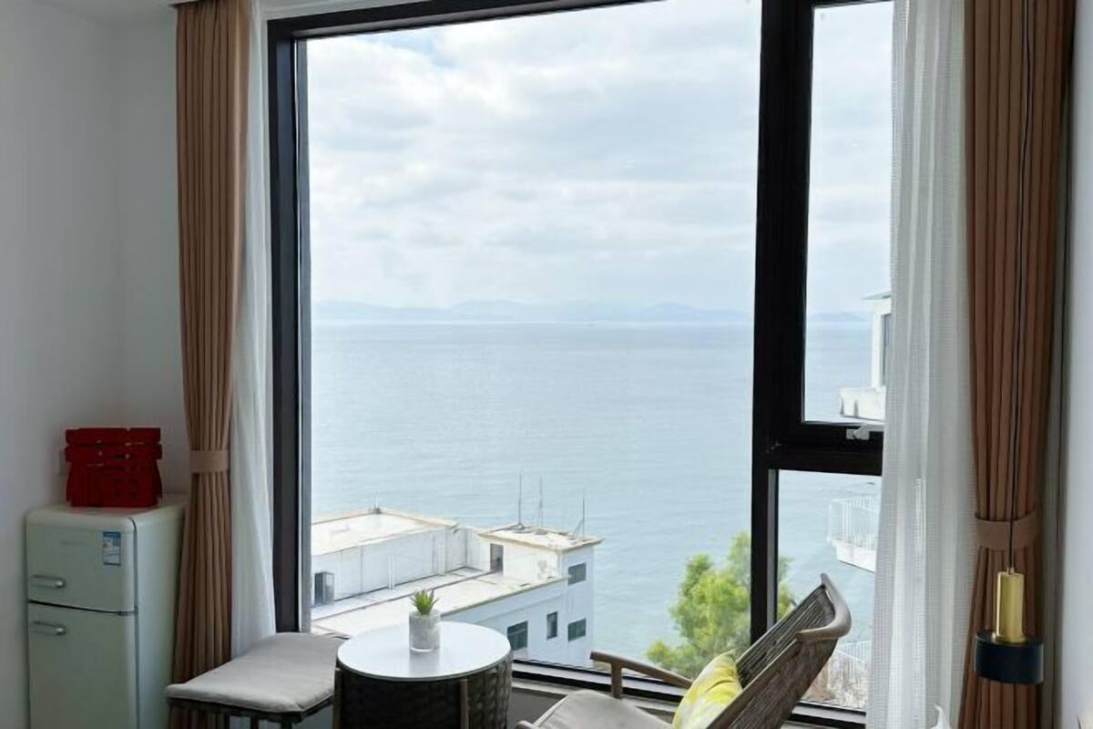 Een kamer met een groot raam, met uitzicht op de zee en verre bergen.