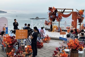 An outdoor wedding ceremony with guests seated on chairs, a stage with floral decorations, and a view of the sea.