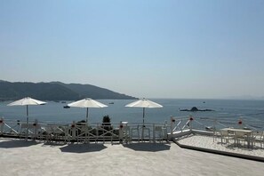 Una terraza con sillas blancas y sombrillas que da a un mar tranquilo con barcos, con montañas al fondo.
