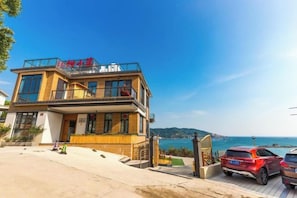 A two-story building with a balcony, a carport with two cars, and a view of the sea.
