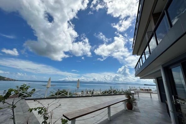 A modern building with a balcony overlooking a body of water, a clear sky, and a few potted plants.