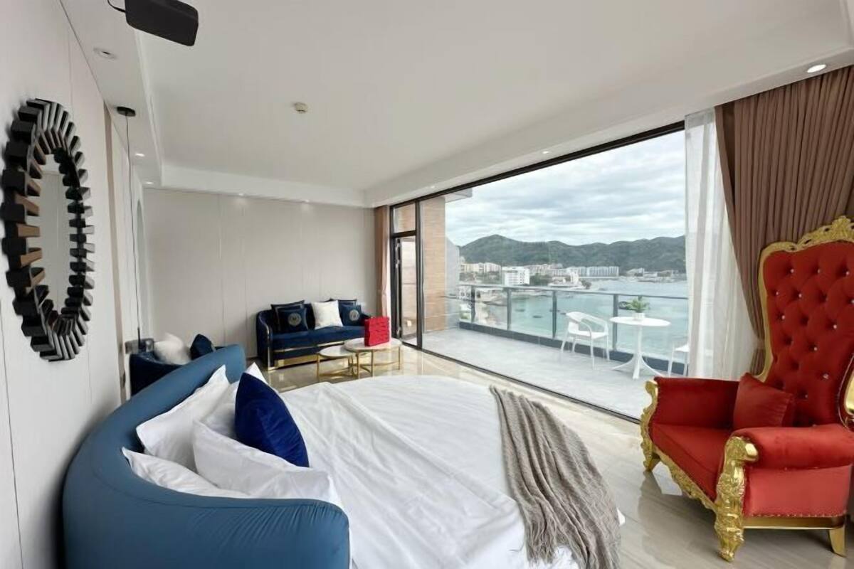 Een moderne hotelkamer met een groot bed, een rode fauteuil, een bank en een balkon met uitzicht op de stad.