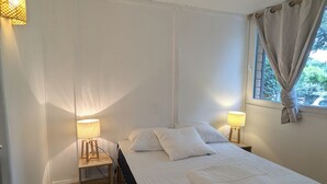 A bedroom with a bed, two bedside tables with lamps, a window with curtains, and wooden flooring.
